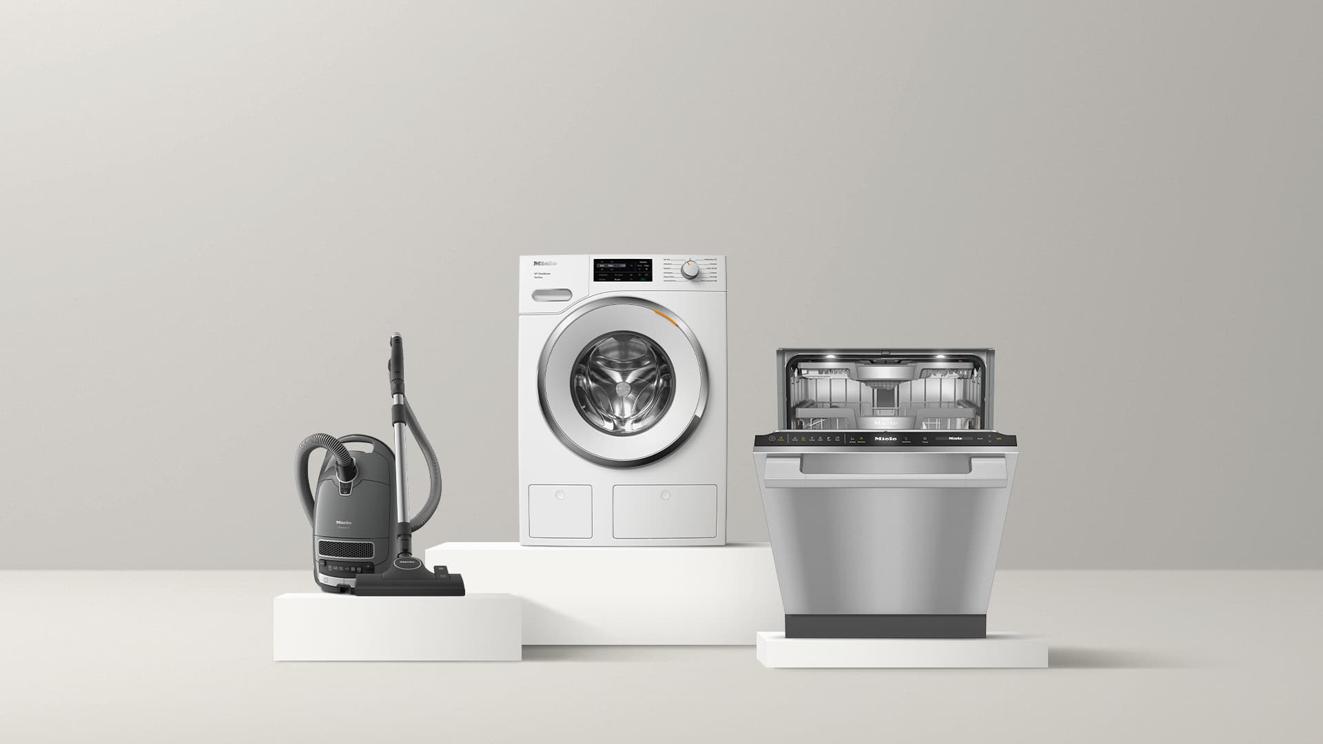A grey canister vacuum cleaner, a white front-loading washing machine, and a stainless steel dishwasher, all placed on pedestals against a grey background.